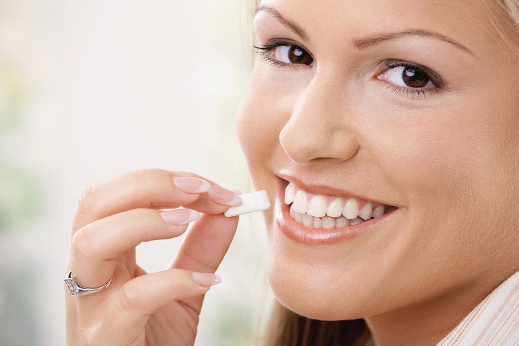 Smiling woman holding a piece of chewing gum near her mouth, showcasing her white teeth.