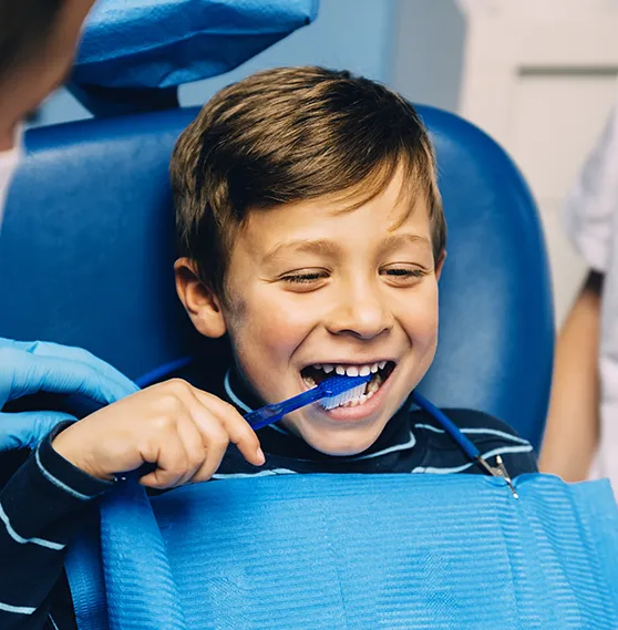Smiling child brushing teeth in a dental chair, assisted by a dental professional.