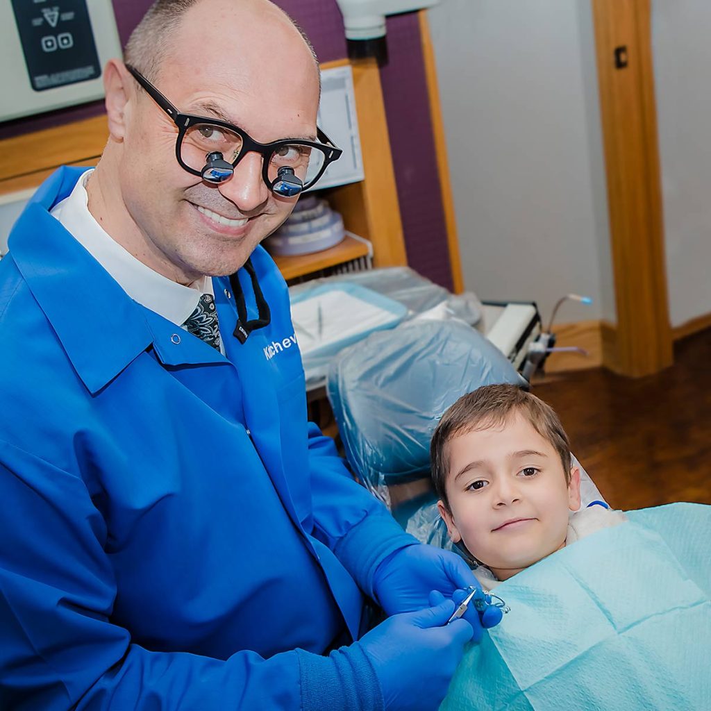 Dr. Boris Kaltchev, a dentist wearing magnification glasses and blue scrubs, smiling while attending to a young boy in a dental chair during an appointment.