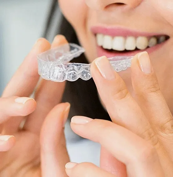 Smiling woman holding a clear dental aligner, preparing to place it on her teeth.