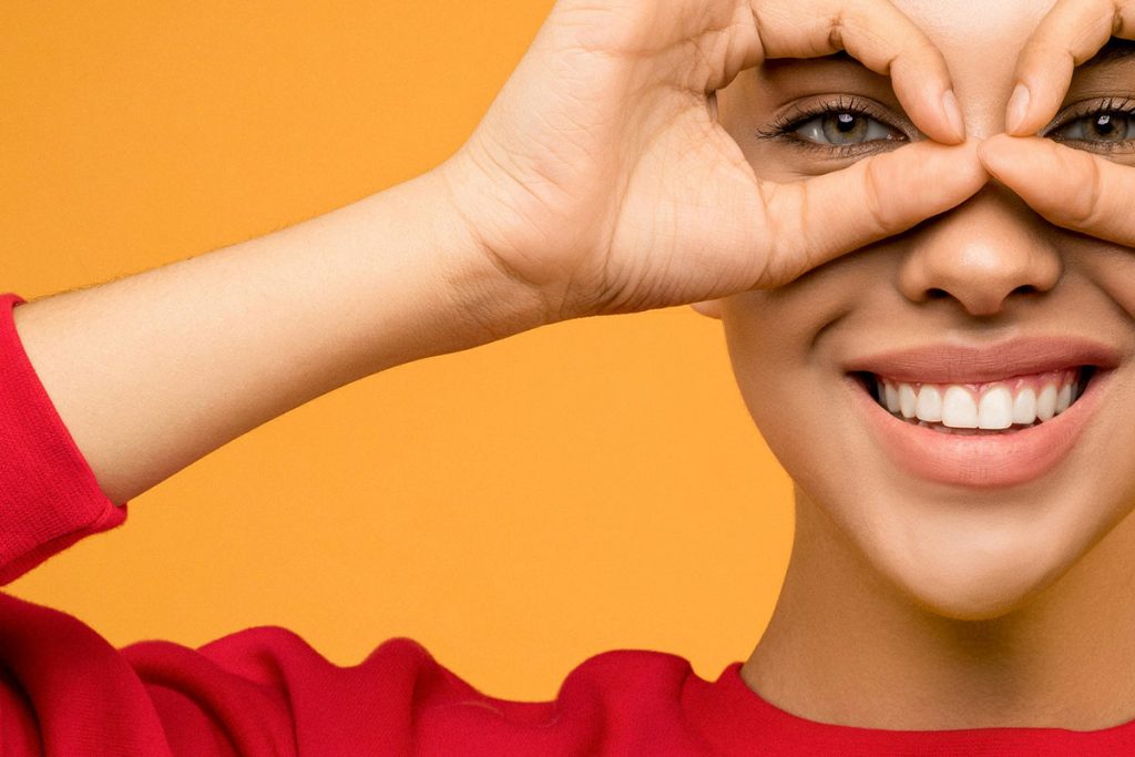 Smiling woman in a red shirt playfully framing her eyes with her hands against an orange background.